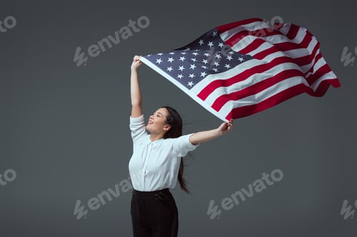 Preview: happy young woman holding american flag in raised hands and looking away isolated on grey