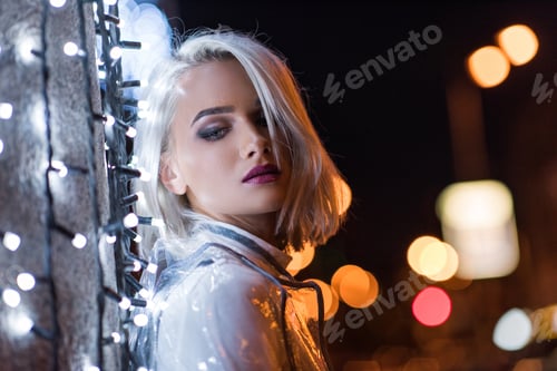 Preview: beautiful young woman leaning on wall with white garland on city street at night