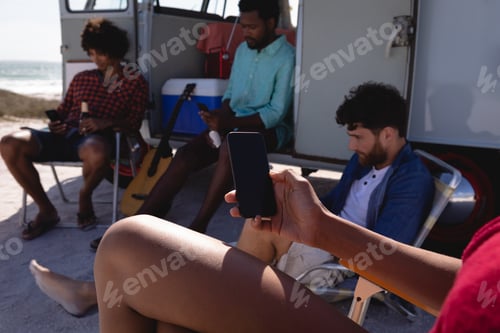 Preview: Group of multi ethnic friends sitting on folded chairs near to a camper van at beach