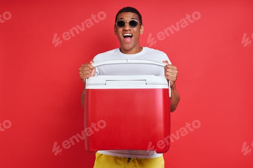 Preview: Happy mixed race man carrying cooler box while standing against red background