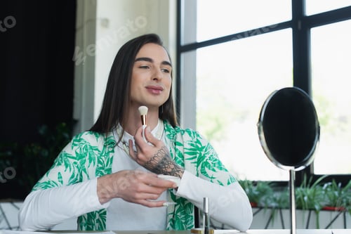 Preview: Smiling queer person holding face powder near mirror in creative agency