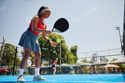 Preview: Black woman serving the ball while playing paddle tennis doubles on the court.
