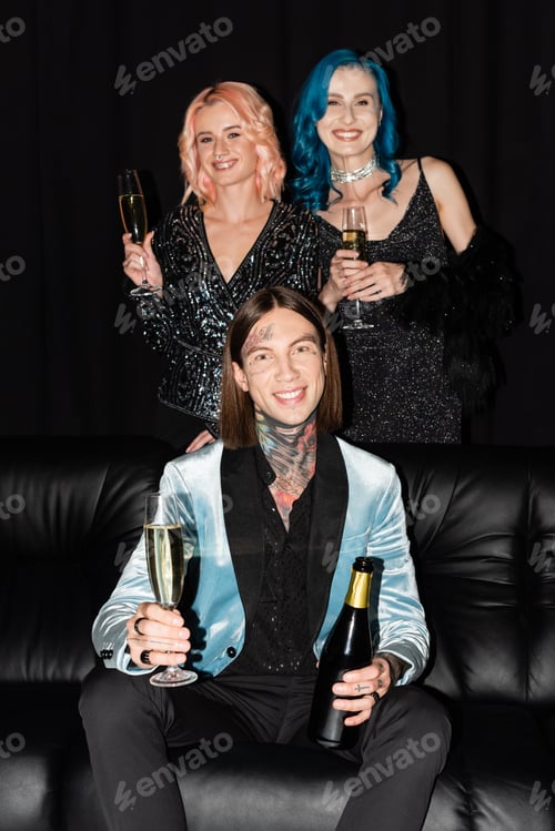 Preview: queer friends holding champagne glasses and smiling near nonbinary person sitting on couch on black