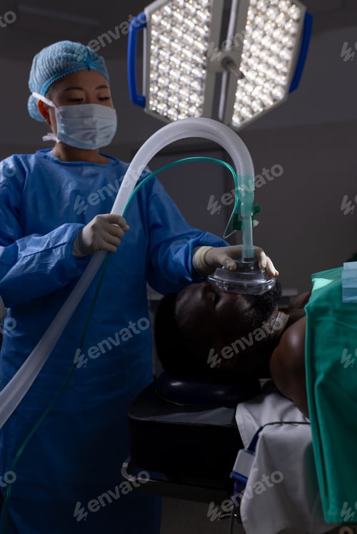 Preview: Asian female doctor with face mask holding oxygen mask on patient face in hospital operating room