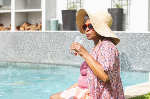 Preview: Senior woman sitting at poolside, wearing straw hat, red sunglasses, sipping from glass, copy space