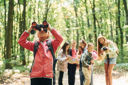 Preview: Girl looking into binoculars. Kids in green forest at summer daytime together