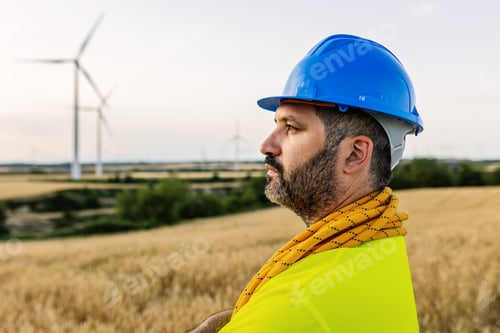 Preview: Male technician worker with blue safety helmet standing in a field of wind farm