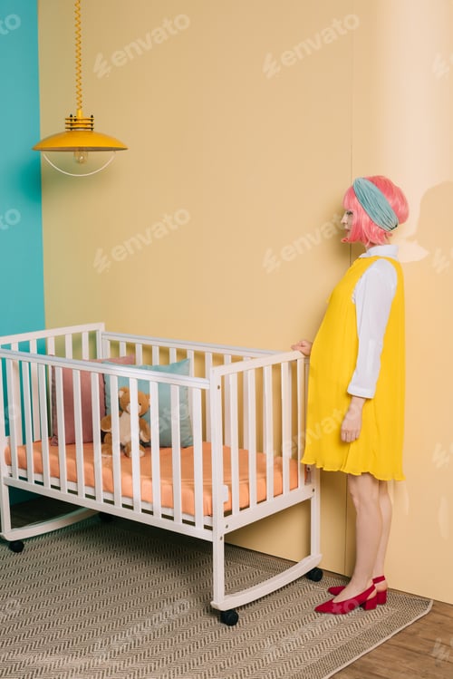 Preview: side view of retro styled pregnant pin up woman with pink hair standing near baby cot in child room