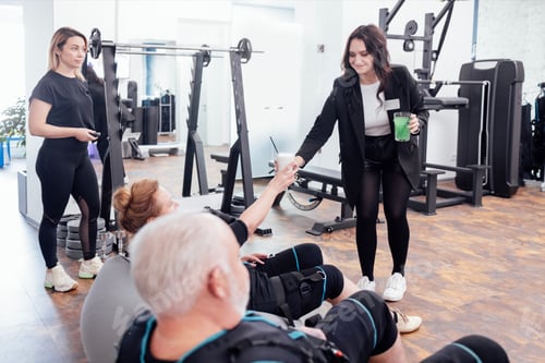 Preview: Smiling female gym worker gives freshly made cocktail to elderly client