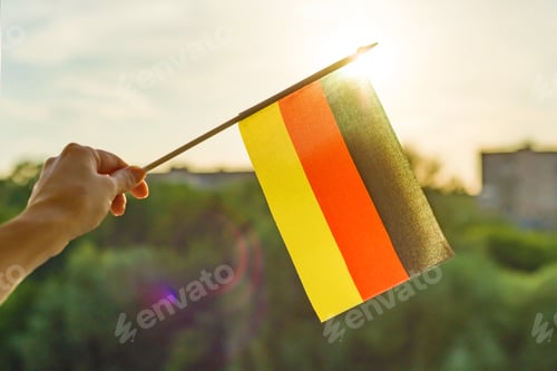 Preview: Hand holds Germany flag an open window. Background blue sky, silhouette of the city, sunset