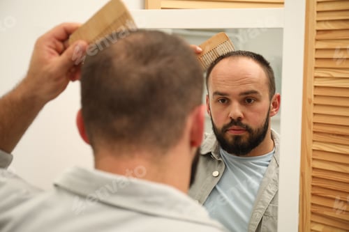 Preview: Baldness problem. Man brushing his hair with comb near mirror at home