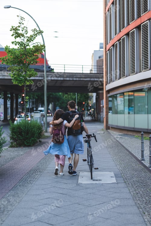 Preview: rear view of couple of tourists with backpacks and bicycle walking at street of Berlin, Germany