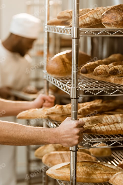 Preview: cropped shot of baker holding shelves with fresh bread loaves at baking manufacture