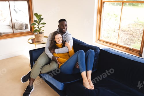 Preview: Happy diverse couple sitting on sofa embracing and smiling at home