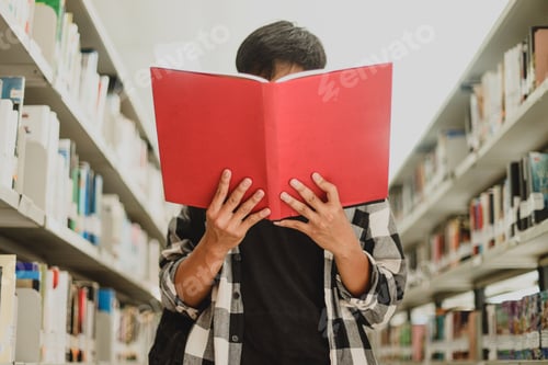 Preview: Young student reading a book and covering his face by bookshelf in the middle of library aisle
