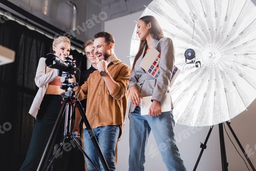 Preview: low angle view of smiling photographer, model, stylist and producer looking at digital camera on