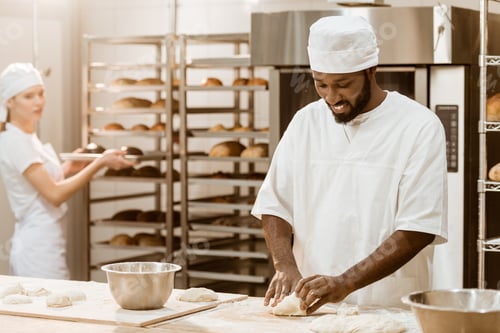 Preview: handsome african american baker kneading dough while his female colleague working on background at