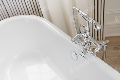 Preview: Shiny metal chrome faucet, elegant water tap on white freestanding bathtub in bathroom