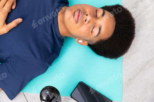 Preview: Biracial man doing yoga lying on mat meditating, using smartphone and earphones