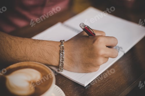 Preview: A coffee cup on wood bar beside window in cafe with hipster man hand writing on notebook.