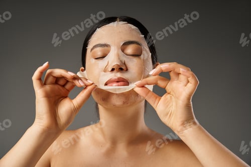 Preview: young asian woman applying hydrating sheet mask for treatment face on grey background, skin care