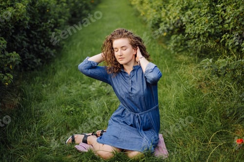 Preview: Happy young woman with brown curly hair, wearing a dress, posing outdoors in a garden