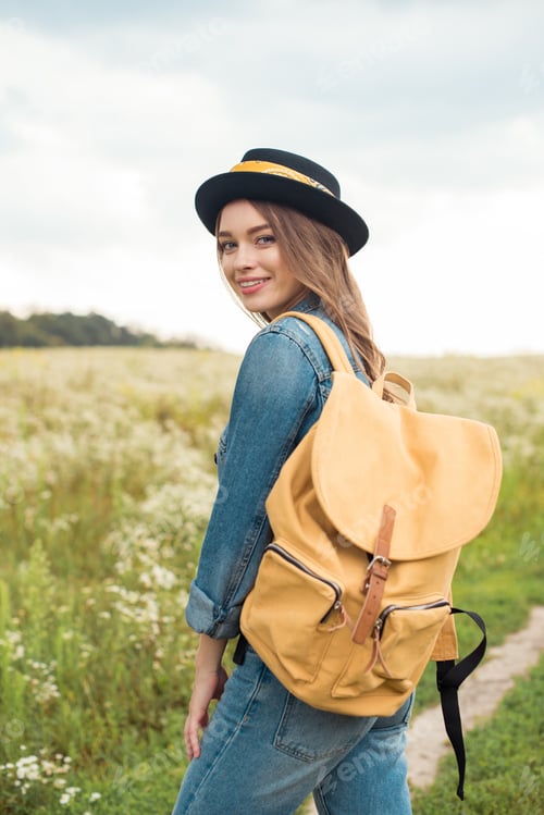 Preview: portrait of smiling attractive woman in hat with yellow backpack standing in field