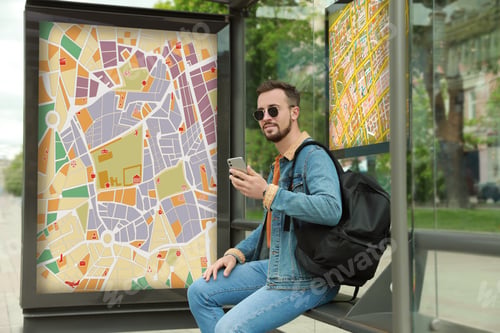 Preview: Young man with smartphone and backpack waiting for public transport at bus stop