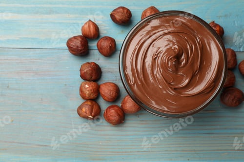 Preview: Chocolate hazelnut spread in bowl and nuts on light blue wooden table, flat lay. Space for text