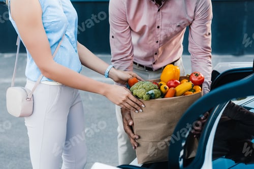 Preview: partial view of couple putting paper bag full of healthy food into car on parking