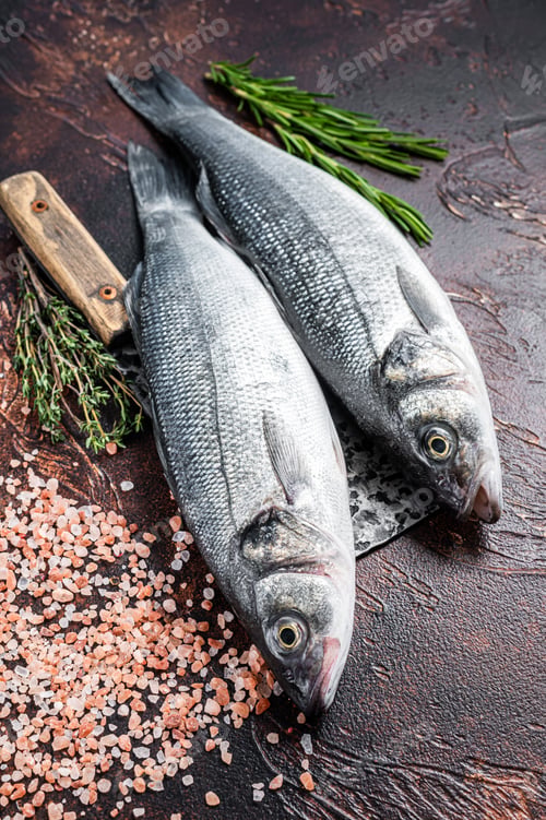 Preview: Seabass or sea bass fresh raw fish on a cleaver with salt and herbs. dark background. Top view