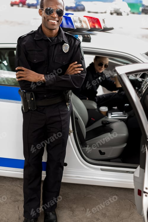 Preview: handsome young policeman with crossed arms standing in front of police car