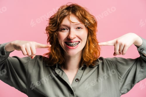 Preview: Portrait of smiling ginger woman pointing fingers on braces on pink background, health care concept