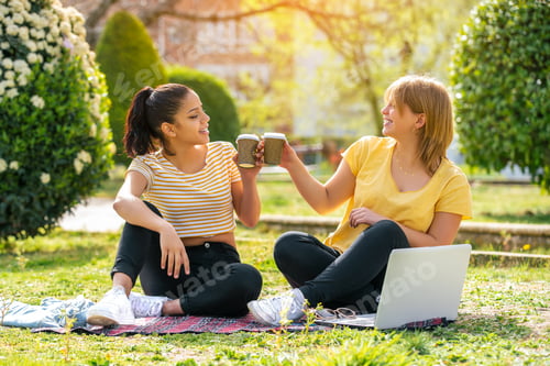 Preview: Two women in the park sitting on a blanket toasting with glasses of coffee on a sunny day