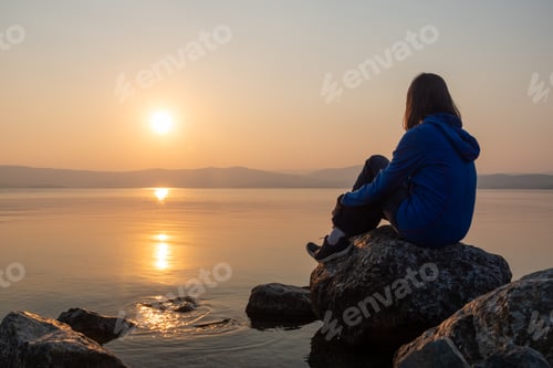 Preview: Girl looking sunset on sea, lake. Woman sitting on rocks. Lonely person. Girl alone outside