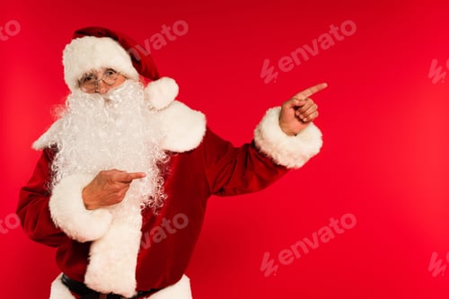 Preview: Santa claus in eyeglasses pointing with fingers and looking at camera isolated on red