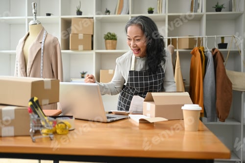 Preview: Happy senior woman small business owner looking at laptop screen and celebrating success