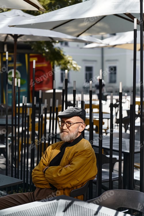 Preview: Dressed in chic attire, an attractive senior man sits with his arms crossed at a contemporary cafe.