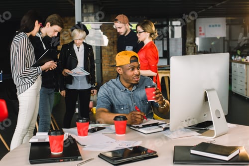 Preview: Smiling young African man drinking coffee while sitting at the table and using computer in modern