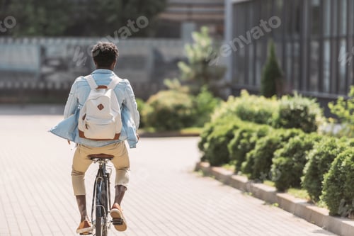 Preview: back view of african american man with backpack cycling along green bushes