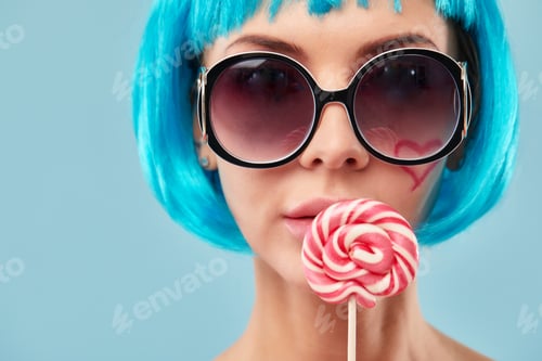 Preview: Young funky girl in blue wig and eyeglasses, holding lollipop candy, looking aside with dreamful