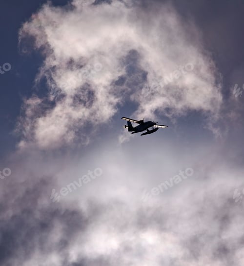 Preview: A small floatplane, a seaplane in the air in a sky full of clouds.