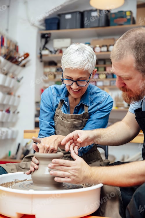 Preview: Senior woman spinning clay on a wheel with teacher at pottery class