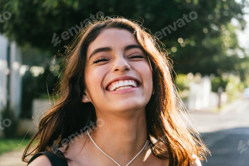 Preview: Delightful ethnic woman with long brown hair smiling and closing eyes on blurred background of