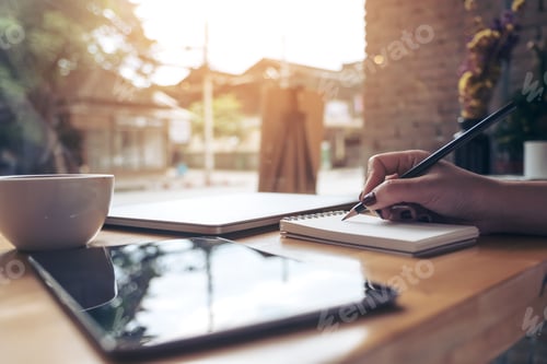 Preview: woman's hand writing on a blank notebook with laptop , tablet and coffee cup on wooden table