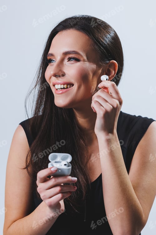Preview: A woman puts wireless headphones into her ears. Female hands hold two white glossy wireless