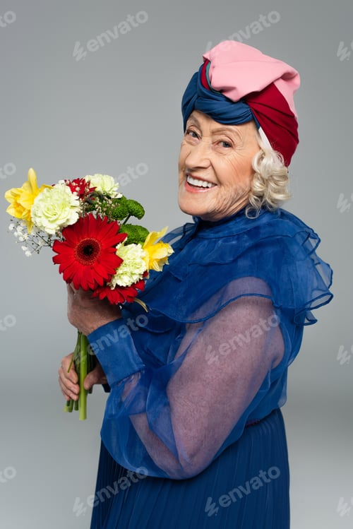 Preview: positive elderly woman in blue dress and turban holding bouquet of flowers isolated on grey