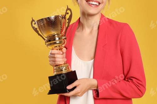 Preview: Young businesswoman with gold trophy cup on yellow background, closeup