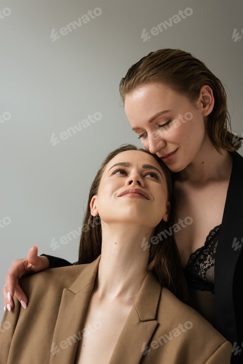 Preview: pretty lesbian woman smiling near seductive lesbian girlfriend in black bra and blazer isolated on