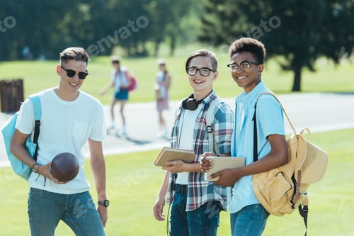 Preview: multiethnic teenage boys with books and backpacks smiling at camera in park